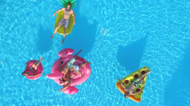 AERIAL TOP DOWN Relaxed girls and guys laying on colourful floaties floating on pool water. Happy smiling friends enjoying summer vacation on inflatable pineapple, pizza, flamingo and doughnut floats