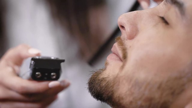 Close up of young bearded man getting haircut on his face by hairdresser at stylish barbershop. Image of female hands using trimmer getting perfect shape for beard and moustache of handsome hipster.