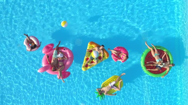AERIAL TOP DOWN: Playful girls and guys laying on colourful floaties in pool. Happy smiling friends enjoying summer vacation on inflatable pineapple, pizza, flamingo, watermelon and doughnut floats