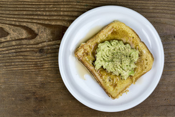 Avocado pear sandwich on hot french toast served on a white plate viewed from above on a wooden table with copy space