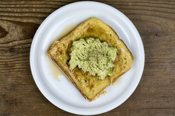 Mashed avocado pear on a slice of toast served on a white plate viewed from overhead on a wood table