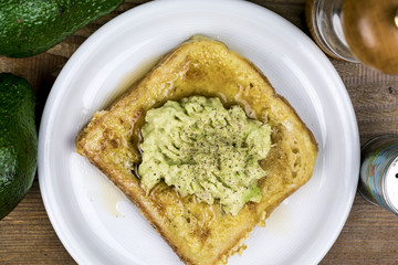 Mashed fresh avocado pear on a slice of toast with salt and pepper seasoning viewed from above on a white plate with ingredients