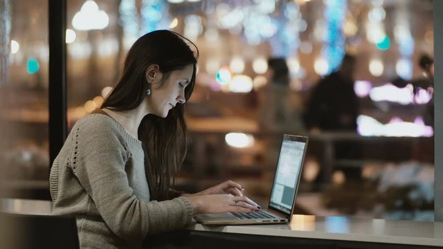 Young beautiful woman sitting near the window in the evening and working on laptop computer. Girl surfing the Internet.