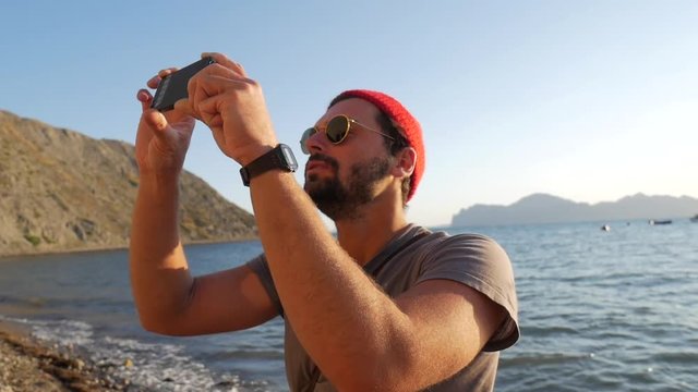 Young Hipster Caucasian Man on a Beach Shooting Video Using Mobile Phone. HD Slowmotion. Crimea, Russia.