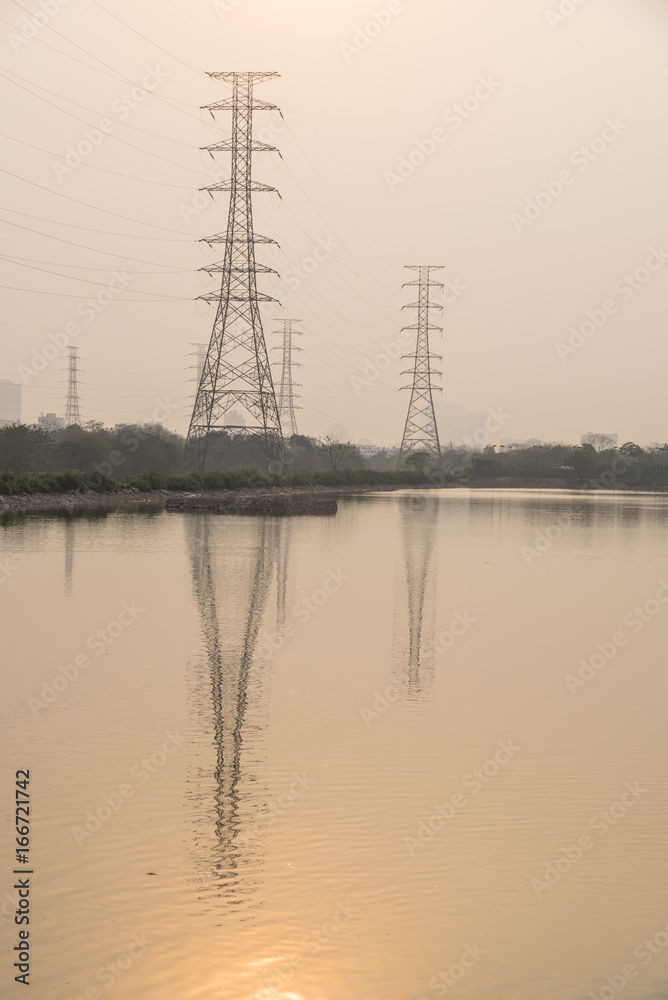 Wall mural Group silhouette of transmission towers (power, electricity pylon, steel lattice) by the lake with sun reflection at sunset in suburb Hanoi, Vietnam. Row of residential building complex in background.