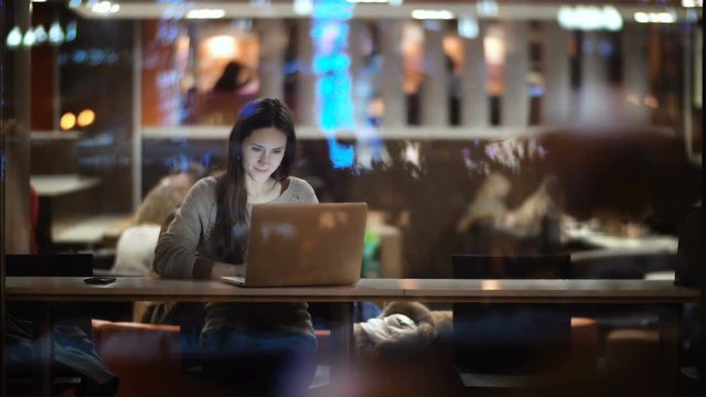 Young beautiful woman sitting at the table and using the laptop. View inside the window on female working in cafe.