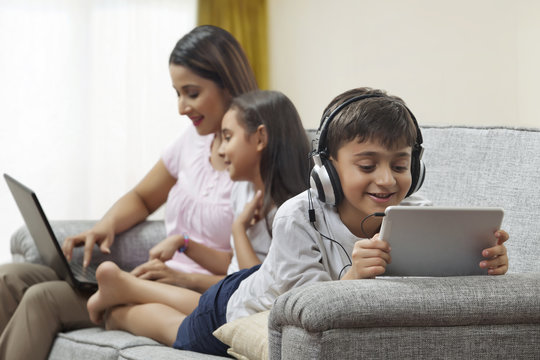 Focused boy using digital tablet headphones with mother and sister using laptop in background