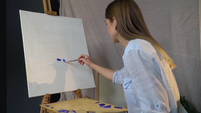Pensive brunette artist makes the first smears of blue paint on a white hulk standing on a wooden easel