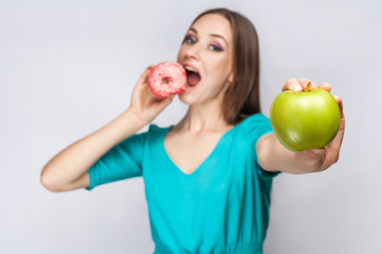 Beautiful young woman with freckles in green dress, eating pink donut and holding green apple. studio shot on light gray background