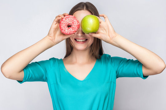 Beautiful young woman with freckles in green dress, holding before her eyes green apple and pink donut with toothy smile. studio shot on light gray background.
