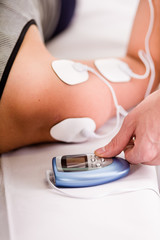 Fototapeta premium Close-up of a personal trainer holding with one hand the electric machine with the electrostimulator electrodes in the shoulder of a deportist, upside down on a bed