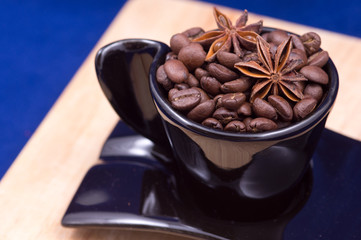 Black cup filled with coffee beans, anise and cinnamon on a wooden surface
