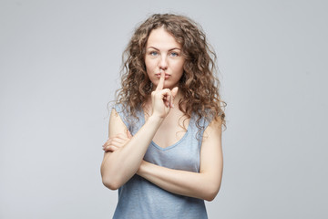 Shh!Attractive Caucasian curly haired lady looking the camera with big blue eyes holding index finger on lips asking to keep silence and not telling her secret. Secrecy, privacy and confidentiality.