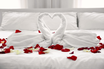 Two towel swans and rose petals on bed in light hotel room