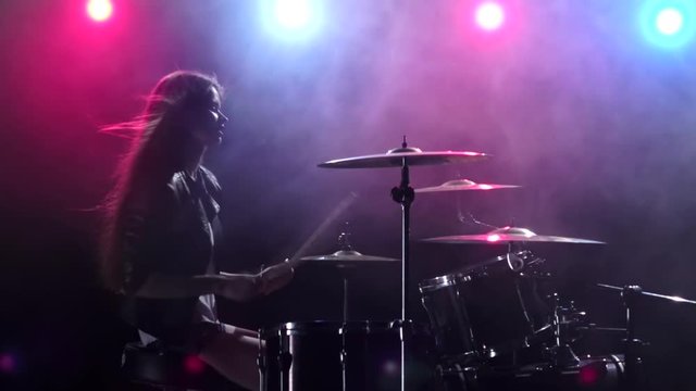 Girl plays the drums and smiles. Black smoke background. Red blue light from behind. Side view. Slow motion