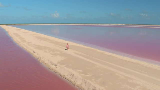 AERIAL: Beautiful young girl on summer vacations walking on sandy levee along gorgeous pink, red and violet saltworks in hot sunny Mexico. In the background stunning turquoise ocean on the horizon