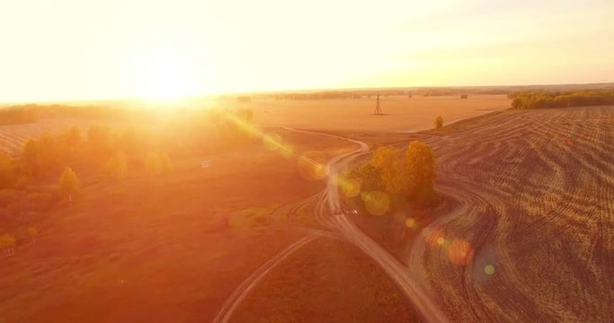UHD 4K aerial view. Mid-air flight over rural dirt road with car. Yellow rural field at sunny autumn evening. Green trees on horizon. Horizontal movement.