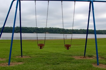 The baby swings at the park with the lake and the overcast skies in background.