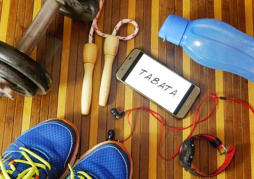 Fitness, healthy and active lifestyles concept. Dumbbells, sport shoes, smart phone with earphone, skipping rope and water bottle gym floor with word tabata