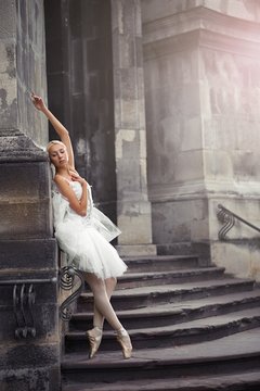 Vertical full length shot of a gorgeous ballerina leaning on the wall of an old castle posing gracefully on the stairs. 