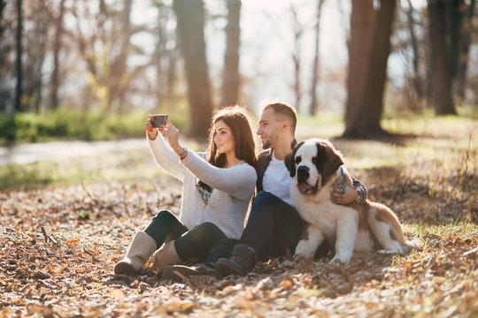Young couple enjoying in park together with their Saint Bernard puppy and taking selfie photo. 