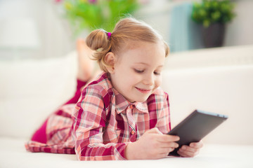 Pretty little child girl with digital tablet, looking and smiling while lying on white couch