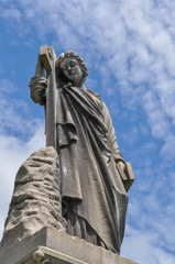 Fototapeta premium Cemetery statue of a person holding onto a book and a cross looking down upon a grave with a blue sky background