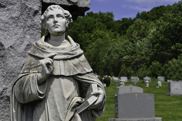 Closeup of a robed scholar statue holding an open book and looking up above a grave in a cemetery