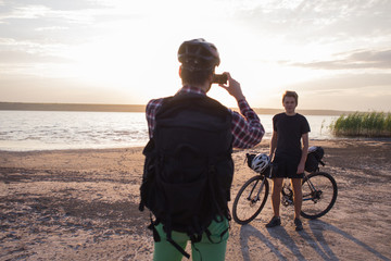 Tourist with backpack make photo of his friend with bicycle during sunrise in lake 