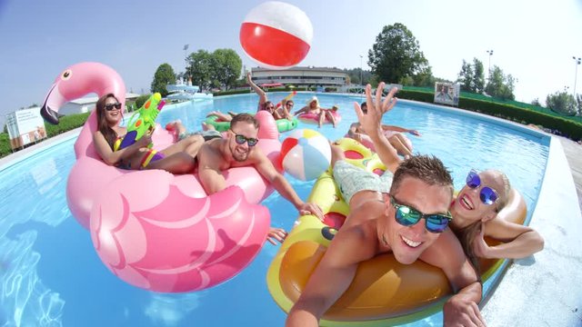 SELFIE CLOSE UP PORTRAIT: Happy smiling friends doing a selfie video on fun colorful floaties in pool. Young people enjoying summer on inflatable flamingo, pizza, watermelon and doughnut floats