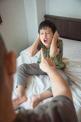 Aggressive asian boy close his ears by hands and shouting to his father on bed at home