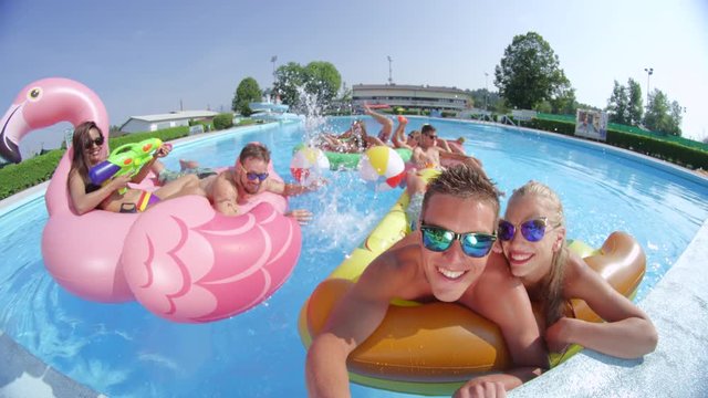 SLOW MOTION SELFIE: Smiling young people enjoying on colorful floaties at pool party Happy teenagers having watergun fight splashing water on inflatable pizza, flamingo, watermelon and doughnut floats