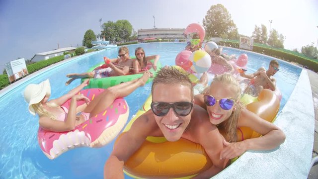 LENS FLARE SELFIE: Smiling young people enjoying on colorful floaties at pool party Happy teenagers having watergun fight splashing water on inflatable pizza, flamingo, watermelon and doughnut floats