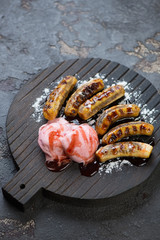 Black wooden serving board with a dessert made of fried mini bananas, ice cream, sauce and powdered sugar, studio shot