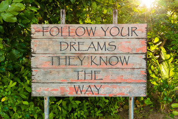 Fototapeta na wymiar Follow Your Dreams They Know The Way motivational quote written on old vintage board sign in the forrest, with sun rays in background.
