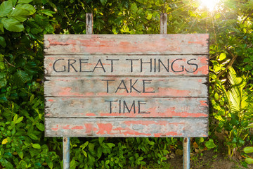 Great Things Take Time motivational quote written on old vintage board sign in the forrest, with sun rays in background.