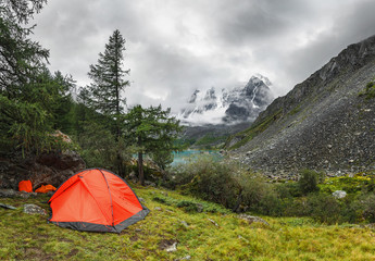 Climbing or hiking camp with a bright orange tent is mounted on a meadow in the mountains of Altai