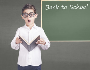 Shocked little geek holding a book in front of a chalkboard with Back to School message