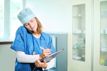 Young female medical doctor in a blue uniform standing in a hospital consulting room, smiling, talking on a mobile phone, looking down