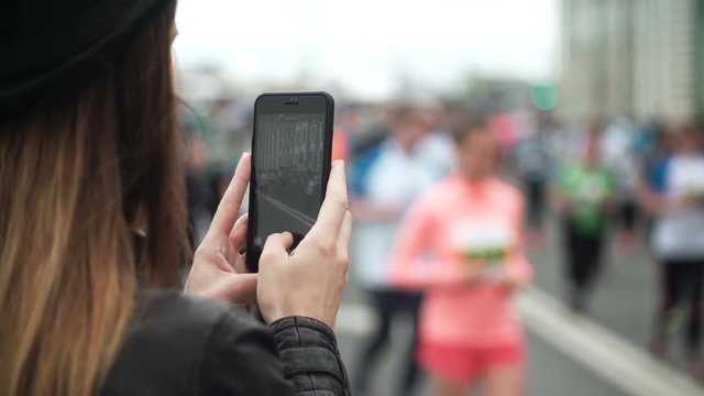 Back view of young woman using the smartphone to filming the video of big group of people running the marathon.