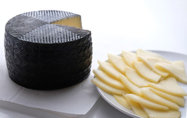 The chopped cheese lies on a plate next to the round head of the cheese. Cheese in black wax lies on a white cutting board