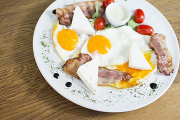 Fried eggs with bacon on the wooden table. Tomato and cheese on the side.