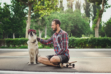 Young hipster man sitting on skateboard with funny siberian husky dog in cap 