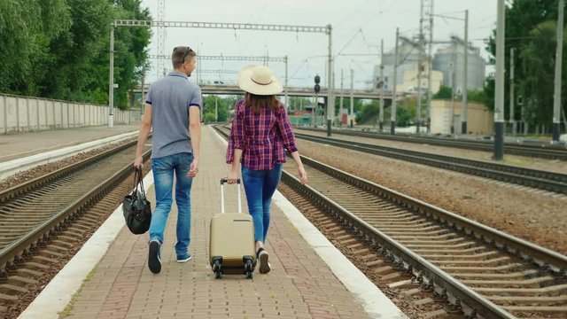 Back view: Young happy couple of tourists with travel bags go along the peron along the railroad. Beginning a great journey. Steadicam shot