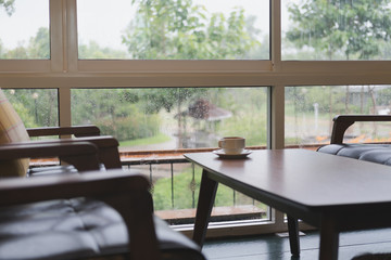 coffee cup on wood table, chair and brown leather sofa near window with garden view at restaurant or cafe in rainy day