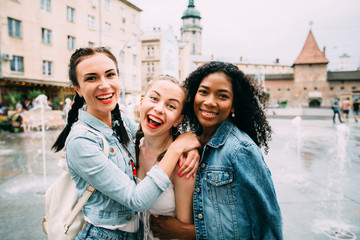 Group of three multiethnic hipster female friends hugging, smiling and looking at camera in summer day over beautiful old city square background.