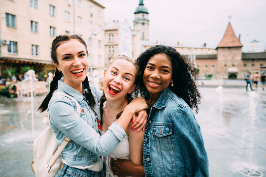 Group Of Three Multiethnic Hipster Female Friends Hugging, Smiling And Looking At Camera In Summer Day Over Beautiful Old City Square Background.