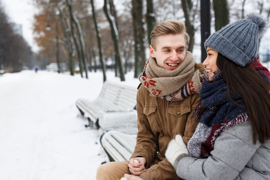Happy guy talking to his girlfriend while sitting on bench in park on wintery day