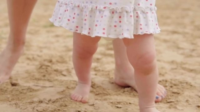 First baby's steps on the beach, mother is holding little girl's hands, slow motion