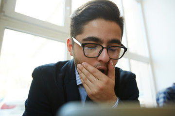 Yawning businessman sitting in front of laptop and shutting his mouth bu hand